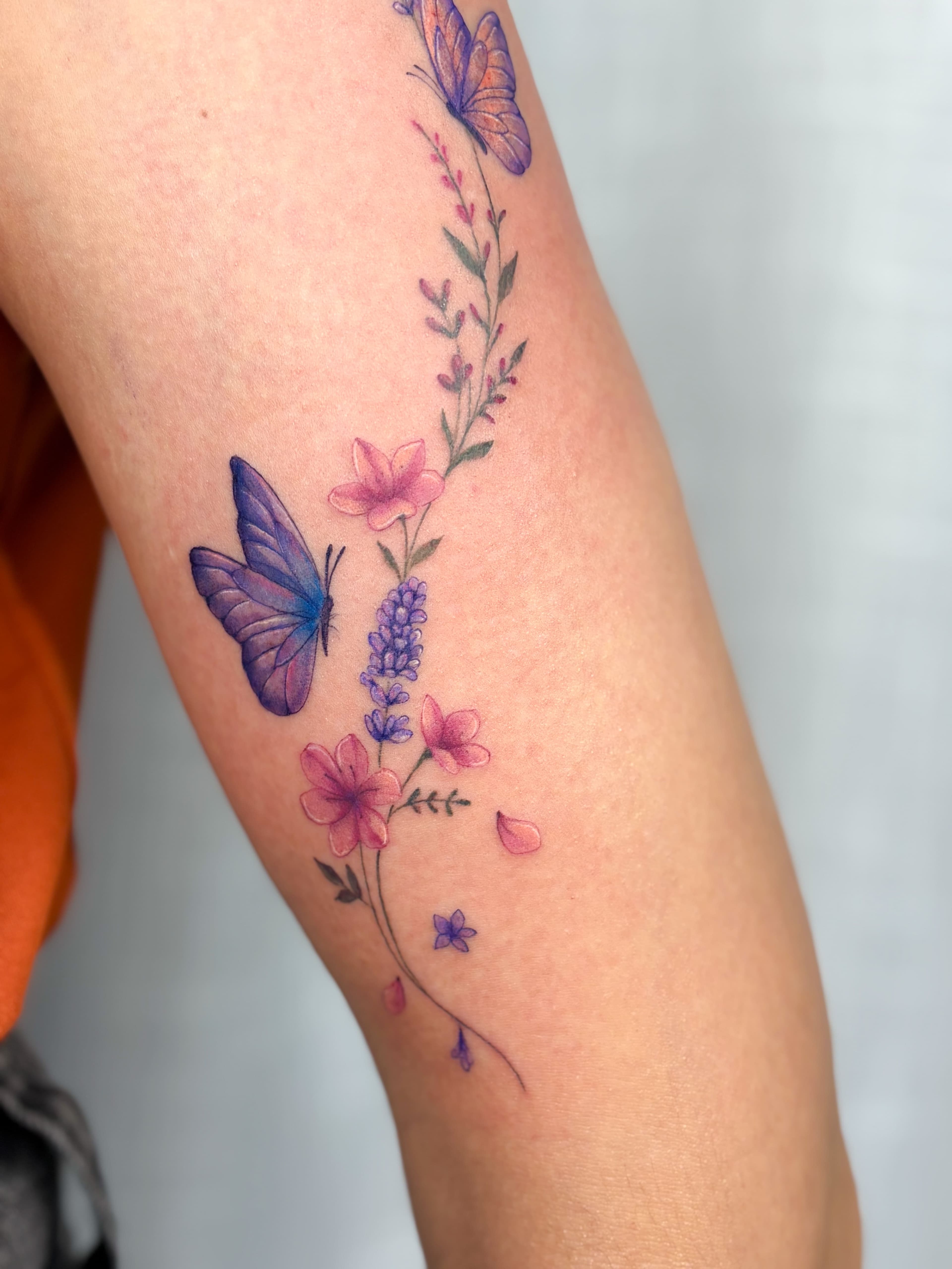 Tatuagem colorida de borboletas e flores em tons de roxo e rosa